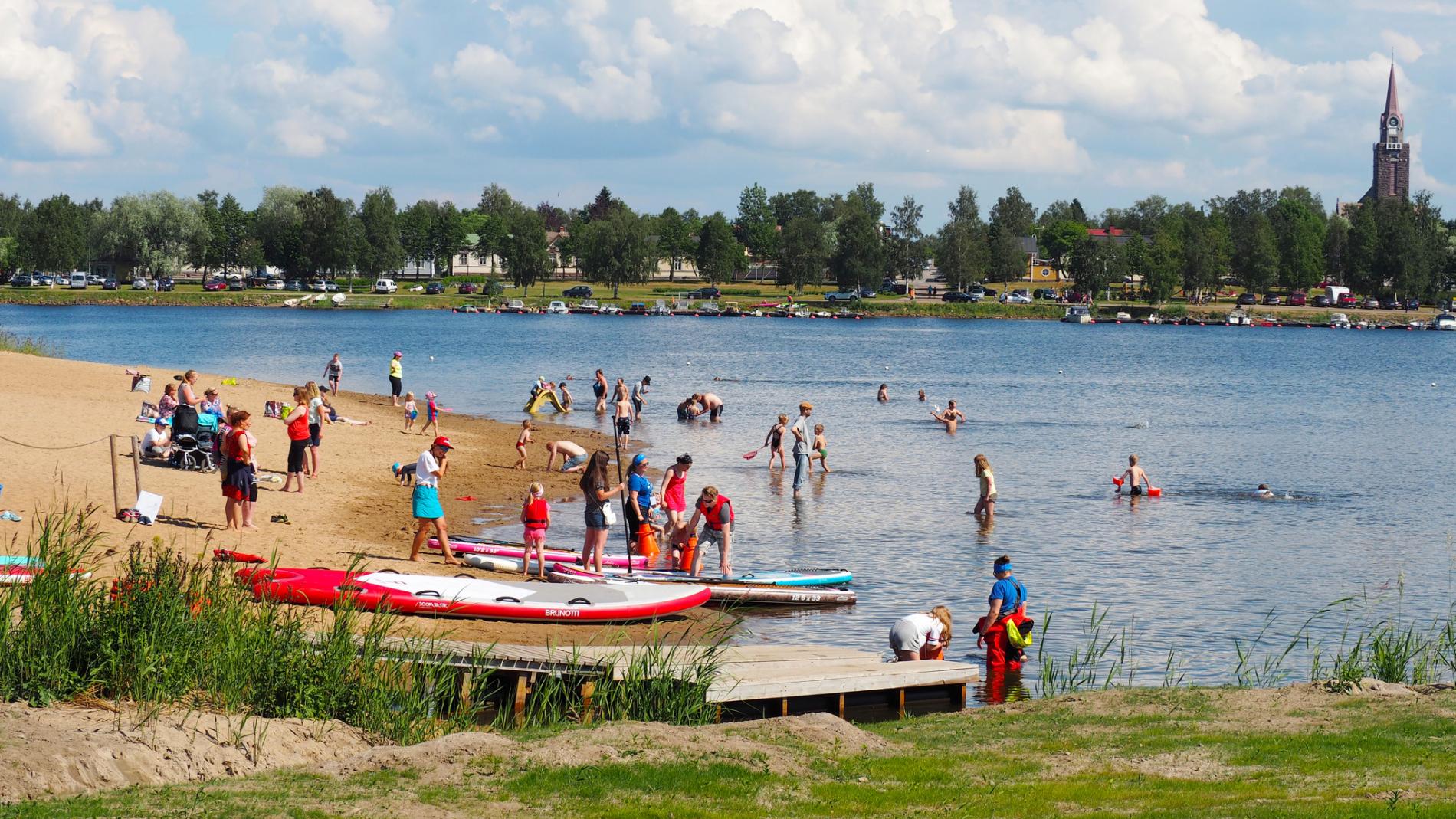 Kesäisellä hiekkarannalla ihmisiä uimassa, osa lähdössä melomaan kanooteilla.