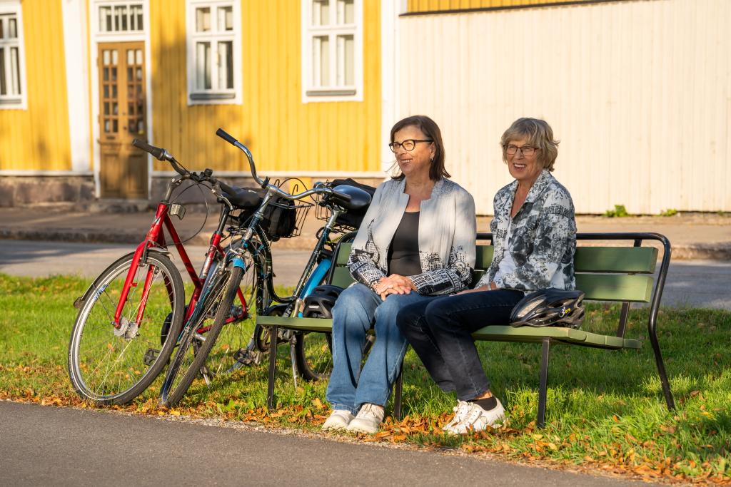 Kuva: Tiina Lämsä. Kaksi polkupyöräilijänaista istumassa penkillä kadun varrella.