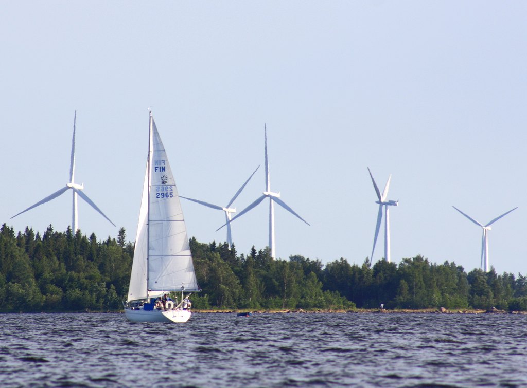 Purjevene merellä. Tuulimyllyjä ja metsää taustalla.
