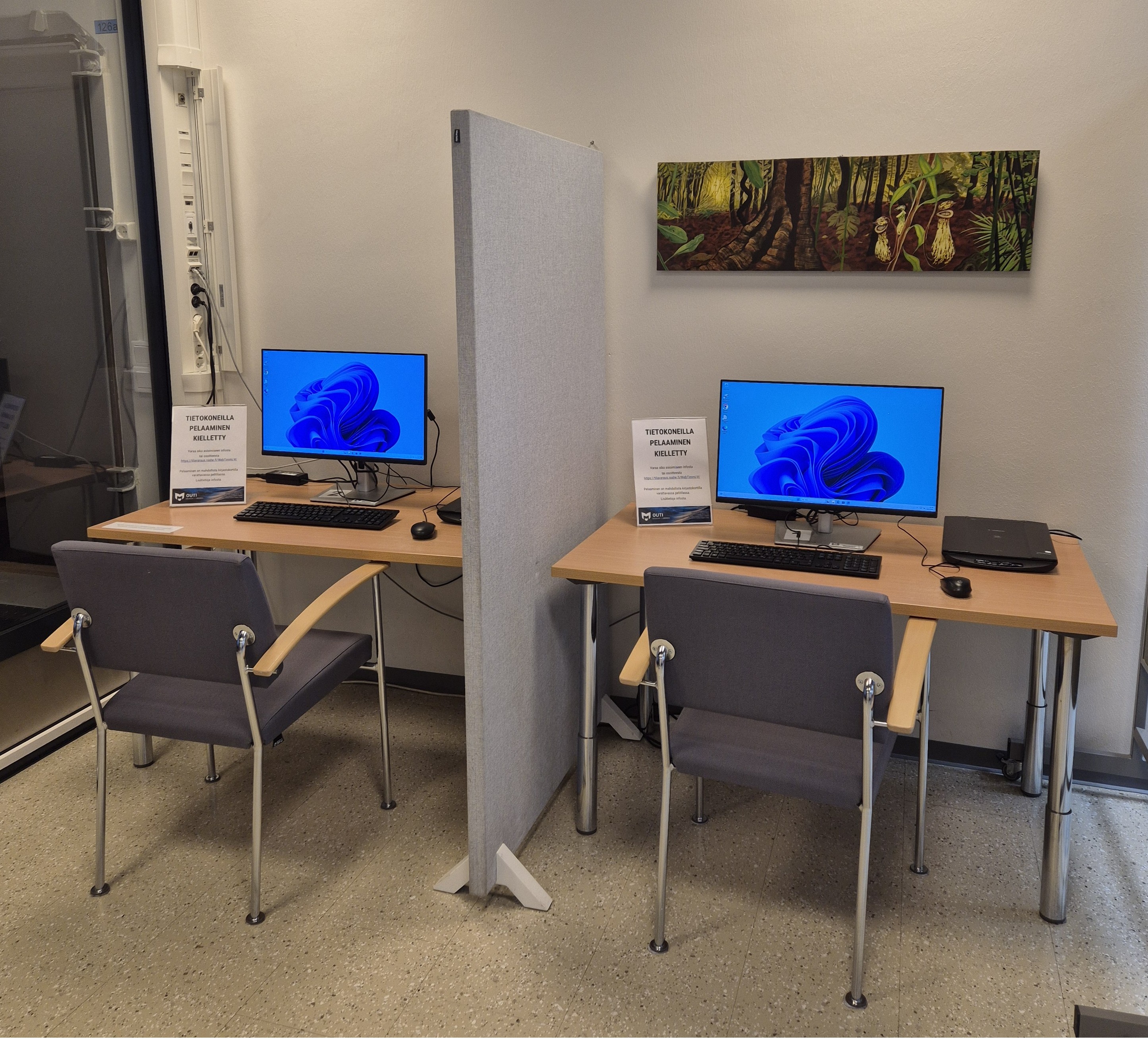 Kaksi asiakastietokonetta seinää vasten, välissään sermi.