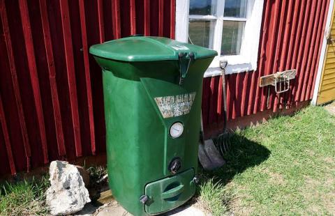 Muovinen vihreä kannellinen kompostori punaisen talon ulkoseinää vasten. 