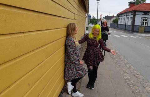 Keltaista seinää vasten kaksi ohjaajaa mekoissa vanhan Raahen kadulla. Yksi ohjaaja pyöräilee kohti huiskuttaen kättä. 