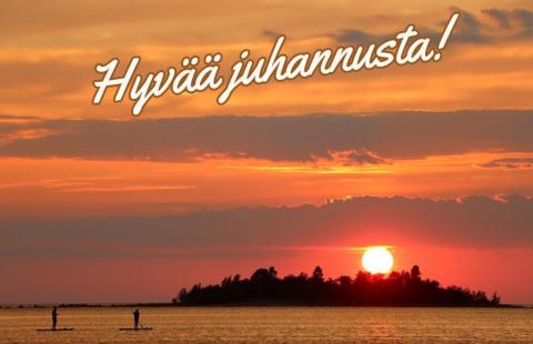 Kaksi ihmistä suppailemassa auringonlaskussa. Kuvan yläosassa teksti, jossa lukee hyvää juhannusta!