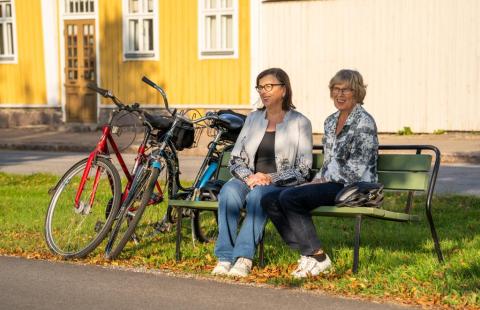 Kaksi polkupyöräilijänaista istumassa penkillä kadun varrella.