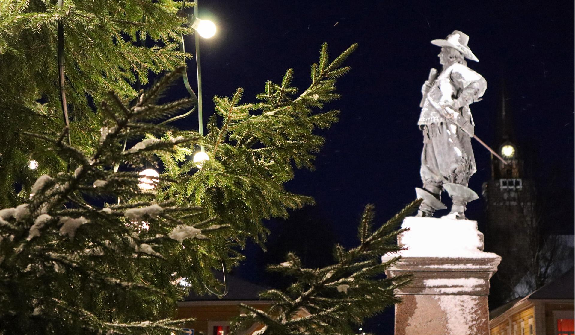 Spruce with Christmas lights. In the background is a statue and a church tower.