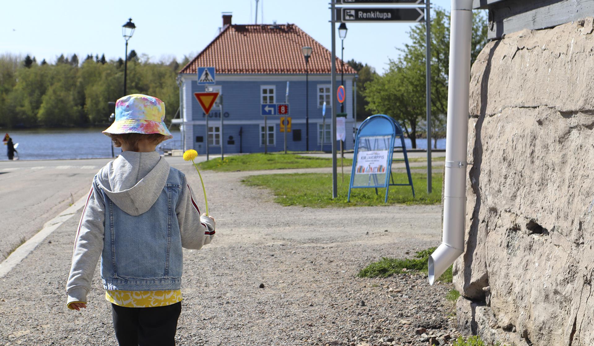 Lapsi, kukka kädessään, kävelee kadulla. Taustalla näkyy meri ja vanha museorakennus.