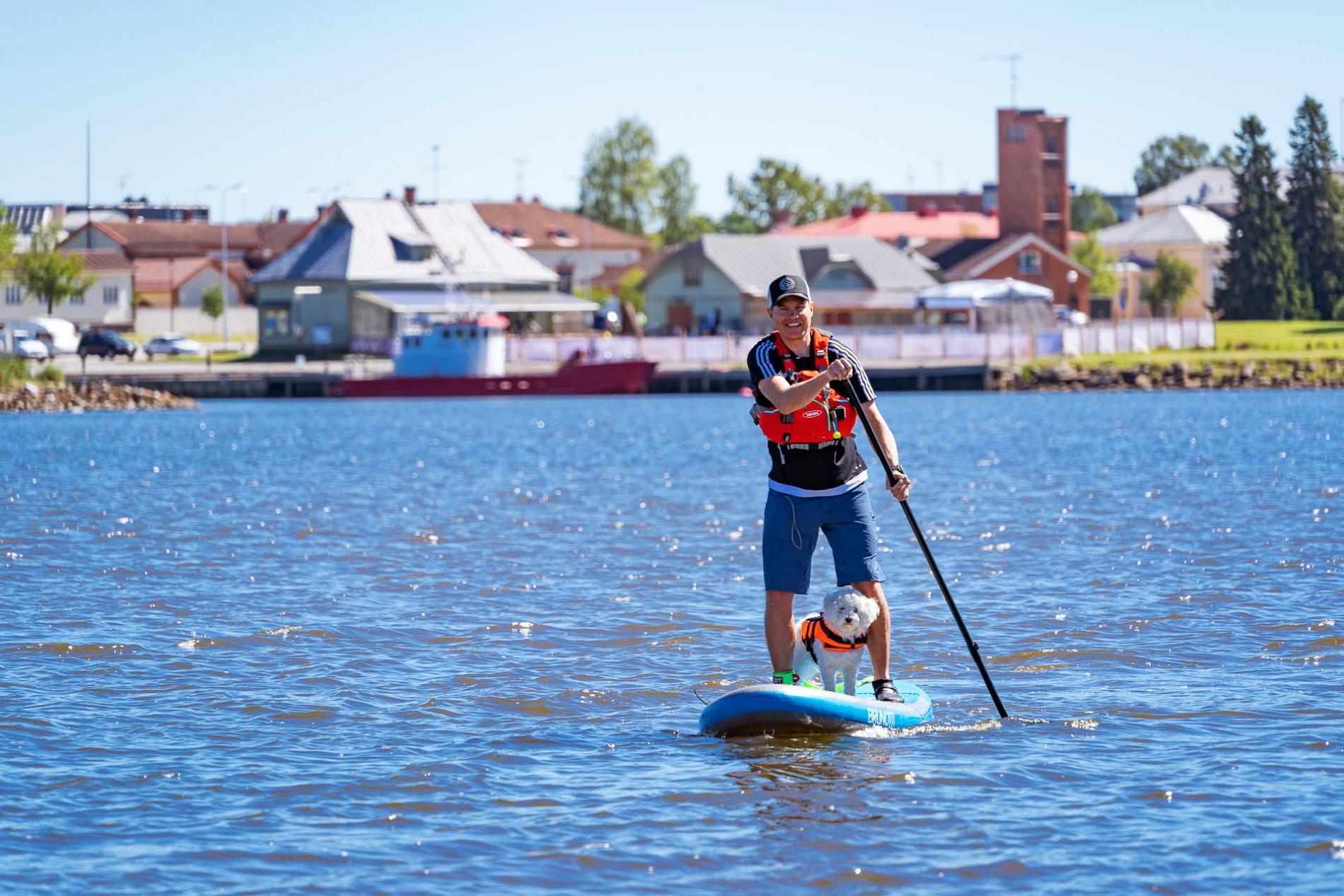 Iloinen mies suppailemassa laudan päällä merellä, pieni koira jalkojen välissä.